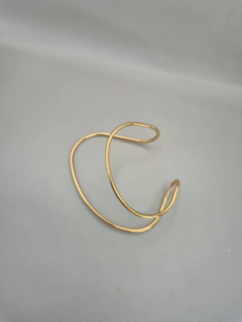 May include: A gold-colored cuff bracelet with a unique, open design. The bracelet features a continuous, flowing shape, creating a modern and minimalist aesthetic. The metal has a polished finish, reflecting light and adding a touch of elegance.