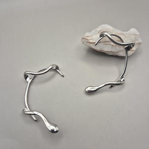 Peut inclure: Une paire de boucles d'oreilles ear cuffs argentées aux formes fluides, lisses et organiques. L'une des boucles est placée à côté d'une petite pierre blanche à la texture rugueuse. Le métal présente une finition polie et réfléchissante.