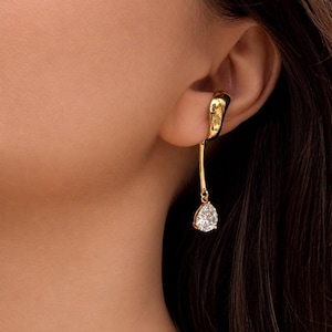 Peut inclure: Une boucle d'oreille en métal doré composée d'une bande supérieure lisse et incurvée qui entoure le cartilage. Une fine tige métallique verticale descend de la boucle, se terminant par une unique pierre transparente en forme de poire.