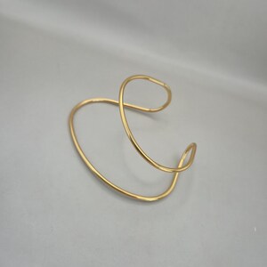 May include: A gold-colored, open cuff bracelet with a flowing, abstract design. The bracelet curves and loops, creating an elegant and modern aesthetic. The jewelry piece is made of a shiny, metallic material.