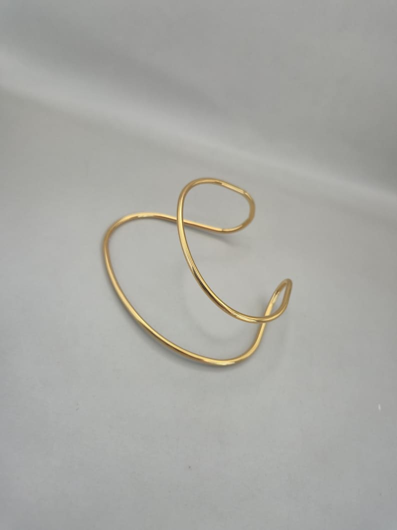 May include: A gold-colored, open cuff bracelet with a flowing, abstract design. The bracelet curves and loops, creating an elegant and modern aesthetic. The jewelry piece is made of a shiny, metallic material.