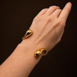 May include: A gold-toned cuff bracelet featuring two polished, teardrop-shaped ends. The open-ended design has a smooth, reflective metallic surface that catches the light, creating a sleek and continuous form around the wrist area.