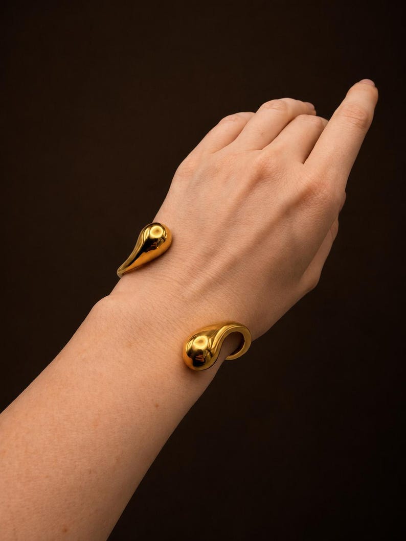 May include: A gold-toned cuff bracelet featuring two polished, teardrop-shaped ends. The open-ended design has a smooth, reflective metallic surface that catches the light, creating a sleek and continuous form around the wrist area.