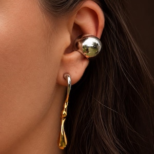 May include: A silver ear cuff and a gold and silver drop earring. The ear cuff is a smooth, rounded shape that sits on the upper ear. The drop earring has a twisted design with a gold teardrop shape.