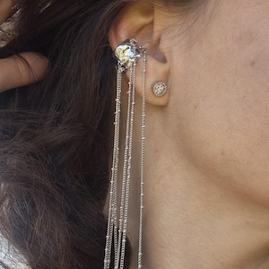 May include: Silver ear cuff with dangling chains and a small stud earring. The ear cuff features a textured design and multiple delicate chains that cascade downwards. The stud earring has a circular design with small, sparkling accents.