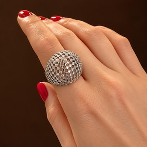 Può includere: Un anello a cupola color argento caratterizzato da un prominente motivo a rete traforata. La superficie dell'anello è ricoperta da una griglia di piccoli ritagli circolari, creando un aspetto sferico e testurizzato sul dito.