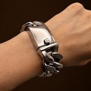 May include: A polished silver-tone metal chain link bracelet featuring a wide, rectangular, smooth-surfaced center clasp mechanism. The bracelet consists of thick, interlocking rounded links that wrap around the wrist, showcasing a reflective finish.