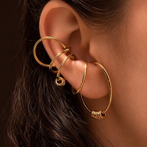 May include: A set of gold-toned ear cuffs and a hoop earring worn on the ear. The jewelry consists of multiple smooth, circular bands that wrap around the cartilage and a larger hoop earring featuring a series of small rings at the bottom.