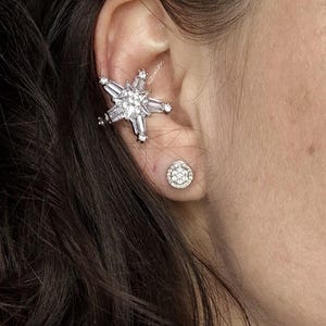 May include: A silver ear cuff and stud earring set. The ear cuff is shaped like a starburst with baguette-cut and round-cut clear stones. The stud earring is a small, round cluster of clear stones.