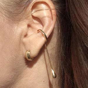 Peut inclure: Une boucle d'oreille ear cuff dorée avec une fine chaîne pendante terminée par une petite breloque lisse en forme de goutte. Une petite créole dorée et épaisse est portée séparément au lobe de l'oreille, complétant le design de l'ear cuff.