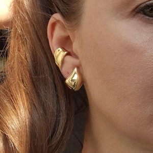 May include: A close-up of a person's ear adorned with a pair of gold-colored ear cuffs. The ear cuffs have a smooth, reflective surface and a unique, curved design. The person's hair is a mix of brown and auburn.