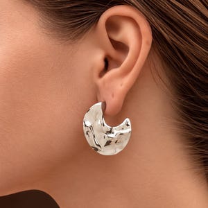 Peut inclure: Une boucle d'oreille argentée en forme de croissant de lune avec une surface texturée, martelée et hautement réfléchissante. La boucle d'oreille présente un design large et incurvé qui repose sur le lobe, avec une finition métallique polie.