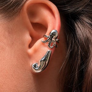 Peut inclure: Une boucle d'oreille en forme de pieuvre aux tentacules courbés reposant sur le cartilage supérieur, accompagnée d'une pièce d'oreille séparée, lisse, allongée et incurvée, positionnée sur le lobe inférieur de l'oreille.