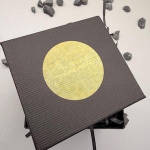 May include: A square, dark brown gift box with a textured, ribbed surface. A circular, translucent yellow sticker is centered on the lid, featuring the text "Bijoux S&eacute;r&eacute;nit&eacute;" in a cursive, light-colored font. A thin ribbon extends from the box.
