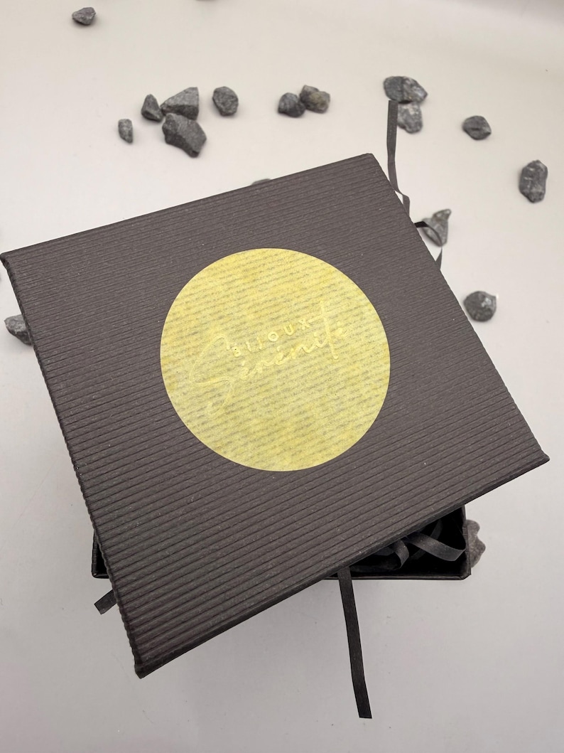 May include: A square, dark brown gift box with a textured, ribbed surface. A circular, translucent yellow sticker is centered on the lid, featuring the text "Bijoux S&eacute;r&eacute;nit&eacute;" in a cursive, light-colored font. A thin ribbon extends from the box.