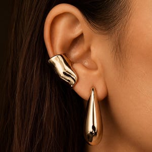 May include: A gold-toned ear cuff with an irregular, molten-like surface texture positioned on the upper ear cartilage, paired with a smooth, polished gold-toned teardrop-shaped earring hanging from the earlobe.