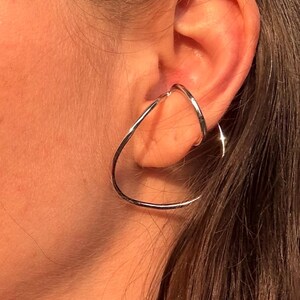 Può includere: Un ear cuff color argento caratterizzato da un design a filo continuo e liscio che forma una goccia irregolare e aperta. La fascia metallica avvolge il bordo esterno della cartilagine dell'orecchio, creando un aspetto lucido e riflettente.