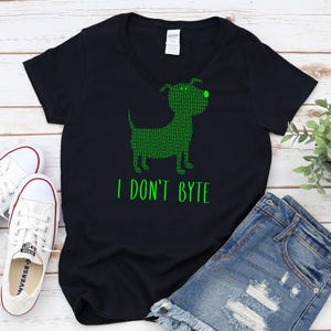 Op de afbeelding: Zwart V-hals T-shirt met een groene hondafbeelding gemaakt van binaire code. De tekst "I DON'T BYTE" staat onder de hond. De afbeelding bevat ook een spijkerbroekshort en witte sneakers.