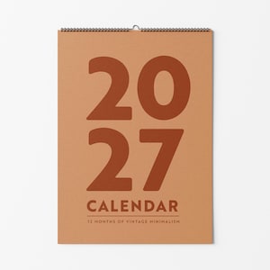 May include: A wall calendar for the year 2027 with a tan background and large, bold, dark orange numbers reading 2027. Below the numbers, the text reads CALENDAR and 12 MONTHS OF VINTAGE MINIMALISM. The calendar is bound with a black wire coil.
