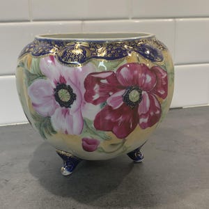 Könnte beinhalten: Ein runder, fußgestützter Keramik-Blumentopf mit handbemaltem Blumendesign. Der Topf zeigt zwei große Mohnblumen in Rosa- und Rottönen vor einem gelben Hintergrund. Der Rand und die Füße sind dunkelblau mit goldenen Akzenten.