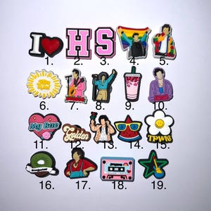 May include: Nineteen colourful shoe charms featuring various designs, including a heart with the word "I", the letters "HS", a rainbow flag, a cartoon figure, a sun, a cartoon figure playing a guitar, a cartoon figure with a microphone, a pink drink, a cartoon figure with a purple shirt, a heart with the words "My Bae", the word "Golden", a cartoon figure with a rose, a watermelon with sunglasses, a daisy with the words "TPW1S", a kiwi, a cartoon figure with a red shirt, a pink cassette tape, and a blue star with a guitar.