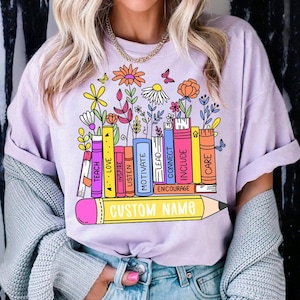 May include: A lilac t-shirt with a vibrant graphic of books, flowers, and butterflies. The books display words such as "TEACH", "LOVE", and "CONNECT". A yellow pencil at the bottom has the text "CUSTOM NAME".
