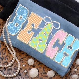 Puede incluir: Una camiseta azul con la palabra "BEACH" en letras coloridas y estampadas. Las letras están delineadas con cuentas doradas y perlas. Un collar de perlas y conchas marinas están dispuestas alrededor de la camiseta.
