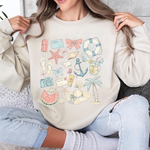 May include: A beige sweatshirt with a summer-themed design. The design features a variety of illustrations, including a watermelon, a seashell, a camera, a butterfly, a life preserver, a starfish, a popsicle, a palm tree, an anchor, and a mason jar with lemonade.
