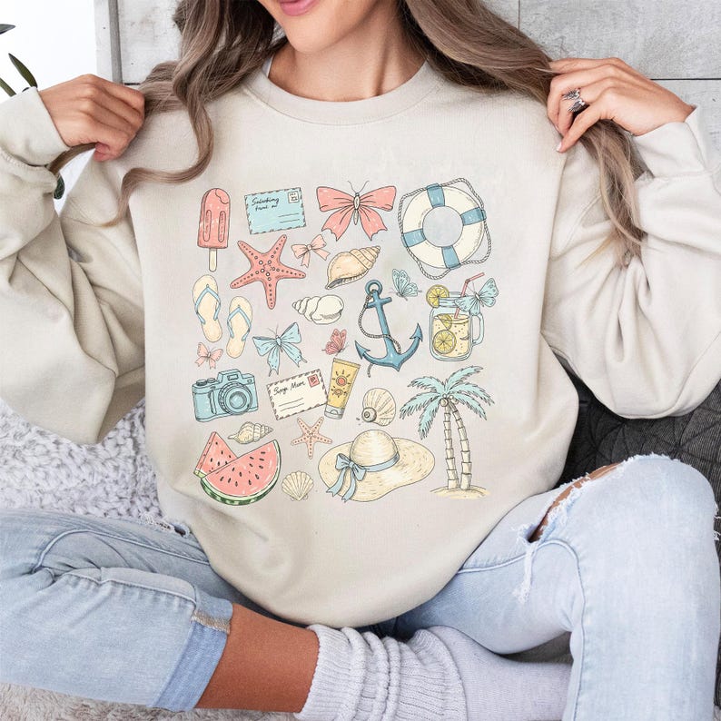 May include: A beige sweatshirt with a summer-themed design. The design features a variety of illustrations, including a watermelon, a seashell, a camera, a butterfly, a life preserver, a starfish, a popsicle, a palm tree, an anchor, and a mason jar with lemonade.
