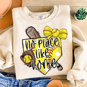 May include: A beige sweatshirt with a softball-themed graphic. The graphic features a softball, a bat, a glove, a bow, and the text "No place like home".