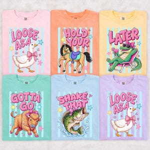 Peut inclure: Six t-shirts aux couleurs pastel avec des graphiques de dessins animés et du texte. Les chemises présentent une oie, un cheval, un alligator, un taureau, un poisson et une autre oie, chacune avec une phrase amusante. Les chemises sont rose clair, orange, jaune, sarcelle, bleu clair et lavande.