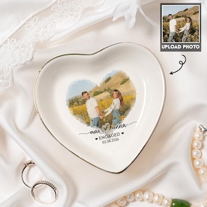 May include: A white heart-shaped ceramic trinket dish with a gold-toned rim. The center features a printed photograph of a couple in a field, with the text "max & anna", "ENGAGED", and the date "02.08.2026" printed in black script and block lettering.