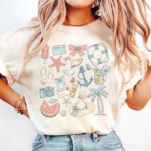 May include: A white t-shirt with a summer-themed design featuring a variety of illustrations, including a watermelon, seashells, a camera, a palm tree, an anchor, a life preserver, butterflies, and a starfish.