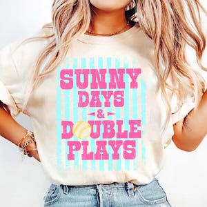 May include: Cream-colored t-shirt with the text "SUNNY DAYS & DOUBLE PLAYS" in pink, set against a light blue striped background. A baseball graphic is featured below the text. The shirt is a casual, everyday wear item.