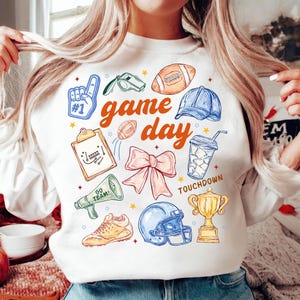 Puede incluir: Sudadera blanca con un diseño colorido de "game day". El diseño incluye un balón de fútbol americano, un casco, un trofeo y otras ilustraciones deportivas. También es visible la palabra "Touchdown".
