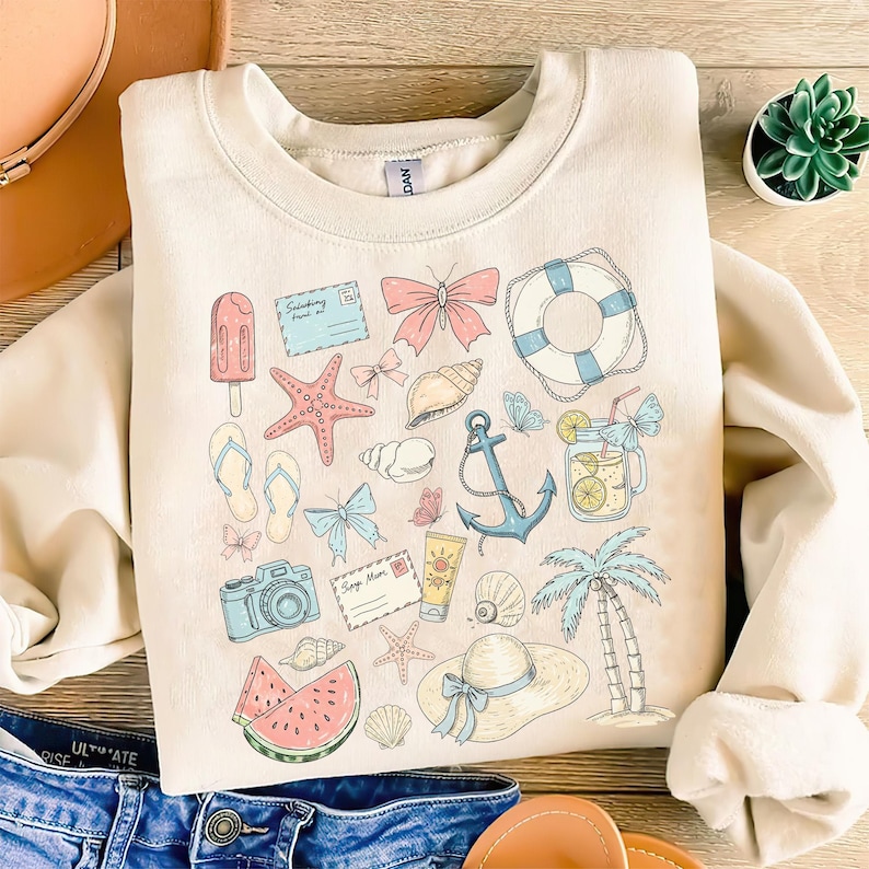 May include: A cream-colored sweatshirt with a summer-themed design. The design features a variety of illustrations, including a starfish, seashells, a camera, a watermelon, a life preserver, a palm tree, and an anchor. The design is in shades of blue, pink, yellow, and green.