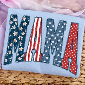 Puede incluir: Una camiseta azul claro con la palabra "MAMA" en letras grandes y decorativas. Las letras están rellenas de varios patrones patrióticos: margaritas, rayas rojas y blancas, estrellas azules y lunares rojos. Los bordes de las letras tienen un ribete festoneado.