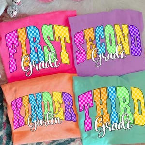 Puede incluir: Cuatro camisetas coloridas con texto de nivel de grado. Las camisetas son rosa, morado, naranja y turquesa. Cada camiseta tiene un nivel de grado diferente: First Grade, Second Grade, Kindergarten y Third Grade. El texto tiene un patrón a cuadros.