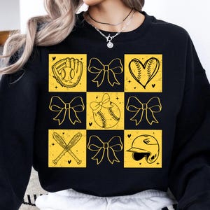 May include: Black sweatshirt with a yellow and black checkered pattern featuring softball-themed graphics, including a baseball, a glove, a heart, bows, and a baseball helmet.