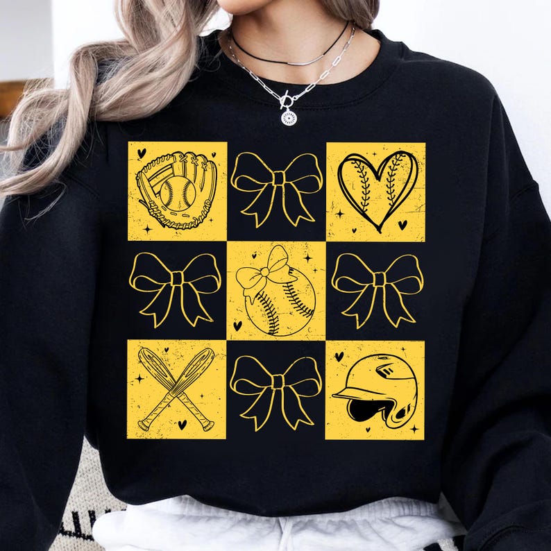 May include: Black sweatshirt with a yellow and black checkered pattern featuring softball-themed graphics, including a baseball, a glove, a heart, bows, and a baseball helmet.