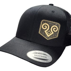 May include: Black trucker hat with a gold embroidered logo of a ram's head with stylized horns. The logo is on a black patch with a detailed pattern. The hat has a black mesh back and a black snap closure. The hat has a label that reads "The Authentic Typoon Snapback Since 1974."