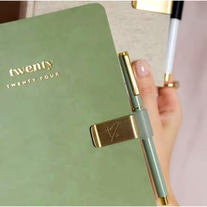 Könnte beinhalten: Ein salbeigrüner Planer mit dem goldenen Text "twenty TWENTY FOUR". Ein goldener Stift wird in einem goldenen Stifthalter gehalten, der am Planer befestigt ist. Der Stift ist salbeigrün mit goldenen Akzenten. Ein weißer und schwarzer Stift ist im Hintergrund.