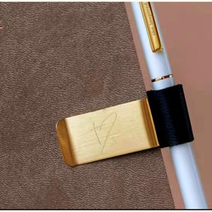 Puede incluir: Un marcapáginas metálico dorado con un diseño en forma de corazón se coloca en un cuaderno de cuero marrón. Un bolígrafo blanco con detalles dorados y el texto "the inspired" se sujeta con un portalápices negro.