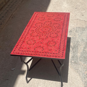 May include: A rectangular table featuring a vibrant red mosaic tile top with an intricate geometric pattern. The tabletop is framed by a decorative border and is supported by four slender, dark-colored metal legs that curve outward at the base.