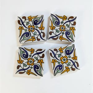 May include: Four square ceramic tiles with a floral design. Each tile features a white background with a repeating pattern of yellow, blue, green, and brown floral elements. The tiles are arranged in a square formation.