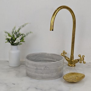 May include: A bathroom scene featuring a gray, cylindrical sink, a gold faucet, and a small, decorative gold soap dish. A white vase with white flowers is on the left. The sink has a diameter of approximately 12 inches.