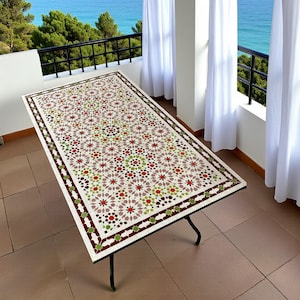 Op de afbeelding: Rechthoekige buitentafel met een wit oppervlak en een kleurrijk geometrisch mozaïekpatroon. Het mozaïek heeft rode, groene en bruine bloemmotieven. De tafel heeft een donker metalen frame en staat op een betegeld terras met uitzicht op zee.