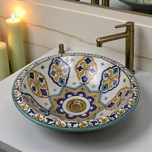 Peut inclure: Un lavabo décoratif en céramique avec un motif peint à la main. Le lavabo présente une base blanche avec des motifs floraux bleus, jaunes et verts. Il est équipé d'un robinet et d'un drain en laiton, et est posé sur un comptoir blanc.