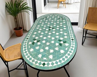 Pode incluir: Mesa de jantar oval com tampo de mosaico em tons de verde-azulado, branco e verde. A mesa tem uma estrutura de metal preto e é rodeada por duas cadeiras de vime amarelas. Uma palmeira em vaso é visível.