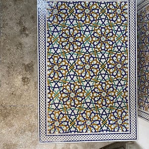 May include: A rectangular mosaic table top featuring a complex geometric pattern in shades of deep blue, mustard yellow, and teal green. The surface is bordered by a white and blue braided tile design, creating a repeating star-like motif.
